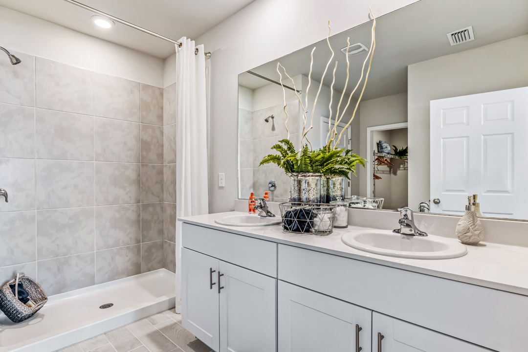 Inside of the Cali model bathroom. Shower-tub combo has grey tiles with a white tub. Dual vanity with white countertops and cabinets with silver hardware. Bathroom also has a closet attached.