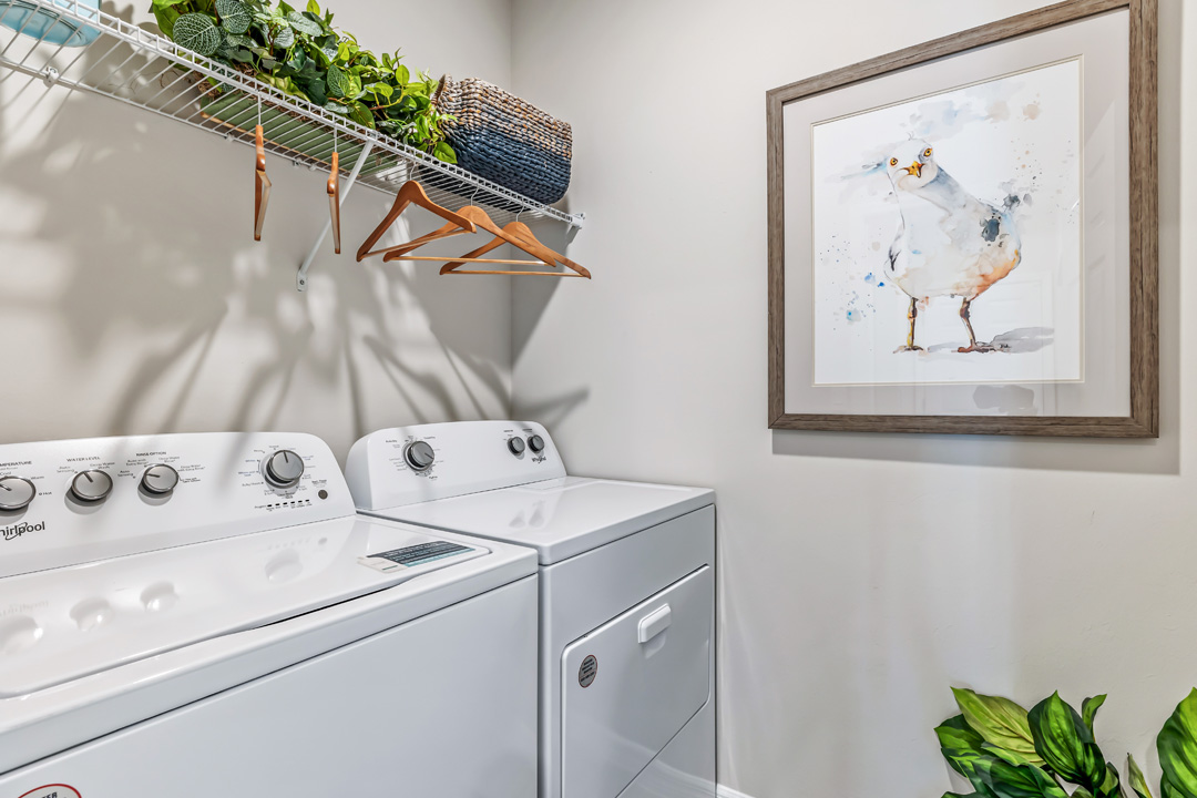 Inside of the Cali model laundry room. Room contains a white washer and dryer side by side. There is also a shelf above the washer and dryer with hangers on it.