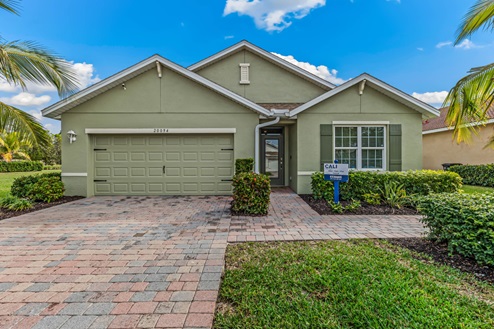 Outside of the Cali model. Green home with two car garage, window with shutter, and front door. Mixed paver driveway.