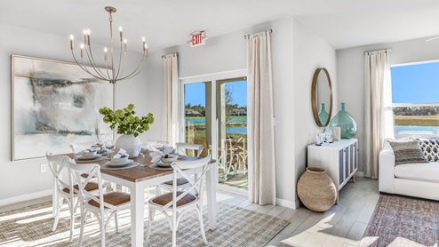The wooden dining table is set for a family of six. There is a chandelier on top and a paitning on the wall behind. There is an added carpet on top of the tile for comfort. The Dining space is well lit from the patio doors. There are curtains on either side of the patio doors.