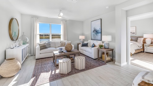 The Living room, there are two white couches, some stools, a coffee table and a chest for storage. There are wooden side tables on either side of the couch. The room is well lit with a window overlooking a view of the back. You can see the entrance to the Master bedroom from here.