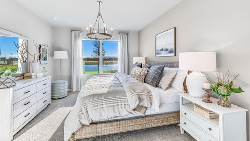 The Master Bedroom is carpeted with grey painted walls. There is a window with a view of the back of the home. There are side tables on either side of the freshly made bed. There is a chandelier above the bed as well. Here you can see the dresser for clothes across from the bed and the lamp in the corner.