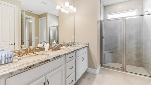 Inside of the Delray B model bathroom. Wood tile flooring with grey walls. Double vanity with grey countertops and white cabinets with silver hardware. Room features a shower with creme tile and glass shower door.