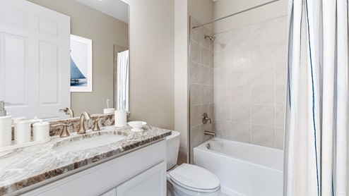 Inside of the Delray B model bathroom. This bathroom has a creme countertop with white cabinets and silver hardware. Shower-tub combo has grey tile and white tub. White toilet between vanity and shower-tub combo.