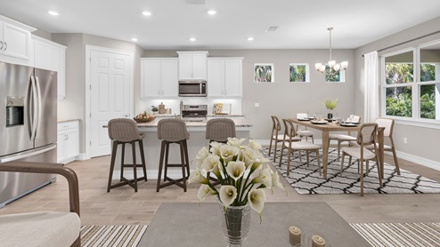 Inside of the Delray B model kitchen and dining area. Kitchen contains an island with bar-style seating, white cabinets with silver hardware, and stainless-steel appliances. Dining area contains a six-person dining table and seats as well as a window and hanging light fixture.