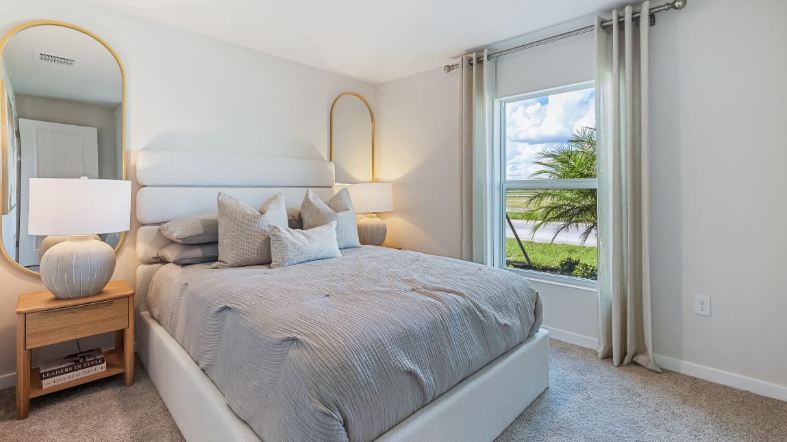 Inside of the Freeport bedroom. White carpet with creme walls. Bedroom has a connected bathroom. Room contains a large bed with grey sheets as well as two small nightstands on either side of bed.
