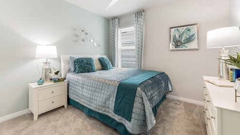 Inside of the Cali model bedroom 1. Crème carpet with one off-white wall and one teal wall. Room contains one bed, a nightstand on one side, and a dresser on the opposing wall. Room also has a window.