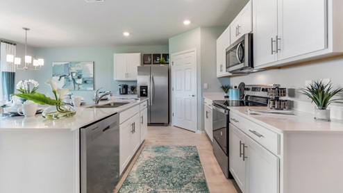 Inside of the Cali model kitchen. Wood tile floors with teal walls. Kitchen contains white countertops and cabinets with silver hardware. There is a kitchen island with bar-style seating. Kitchen appliances are stainless-steel.