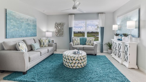 Inside of the Cali model living area. Wood tile flooring and teal walls. Living area contains two couches, an ottoman, storage shelf, and small table. Room also contains a window.