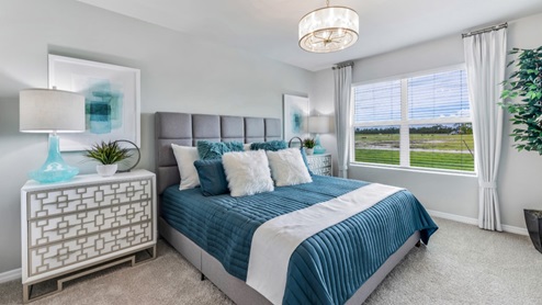Inside of the Cali model bedroom. Crème carpet with off-white walls. Room contains a bed with nightstands on either side as well as a plant in the corner of the room.