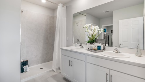 Inside of the Cali model bathroom. Shower has grey tile and silver hardware. Room contains a dual vanity with white countertops and cabinets with silver hardware. Bathroom also contains a closet.