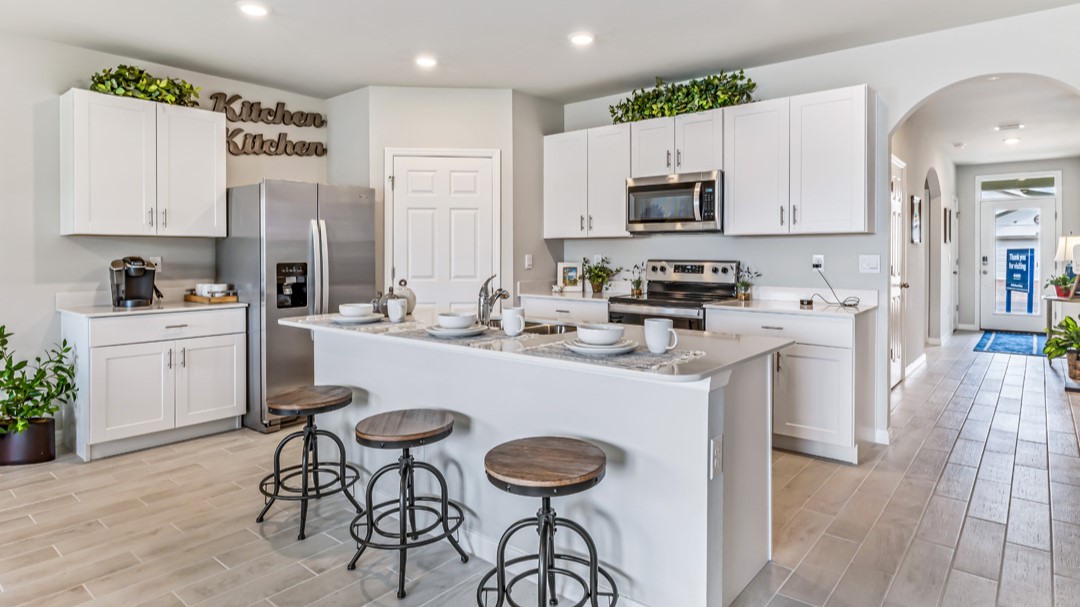 Inside of the Cali model kitchen. Grey wood tile flooring and off-white walls. Kitchen has white cabinets with silver hardware, white countertops, and stainless-steel appliances. There is also a kitchen island with bar-style seating. In the background there is the hallway that connects to the front door and other rooms.