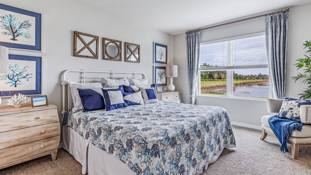 Inside of the Cali model bedroom one. Creme carpet with off-white walls. Room contains a large bed with dressers on either side. There is an armchair in the corner next to the room's window.