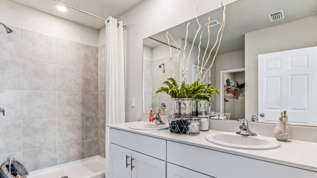 Inside of the Cali model bathroom. Shower-tub combo has grey tiles with a white tub. Dual vanity with white countertops and cabinets with silver hardware. Bathroom also has a closet attached.