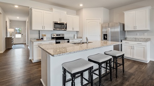 cali plan model in twin lakes community kitchen with granite countertops, bar seating at the island, white cabinets, stainless steel appliances, and a corner walk in pantry