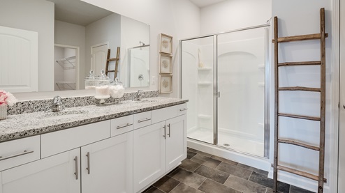 cali plan model in twin lakes community primary bathroom with vinyl flooring, large mirror over a double vanity that has granite countertops, and a walk-in shower with a glass enclosure