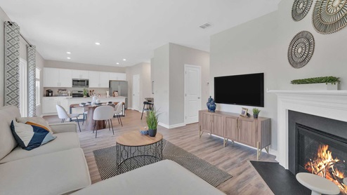 A spacious living area with light wood flooring, neutral-colored walls, and a ceiling fan with lights in the center of the room. The space is bright and airy, with natural light streaming in from large windows on one side.