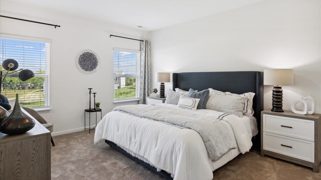 Bright and airy bedroom featuring a large upholstered bed with layered pillows, matching nightstands with modern lamps, neutral decor, carpeted flooring, and two large windows bringing in natural light.