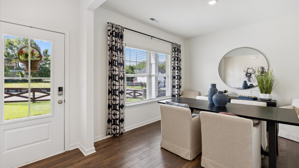 Bright and welcoming dining room with a black rectangular table, upholstered chairs, floral-patterned curtains, a round mirror, and a door leading to the front yard visible through a large window.