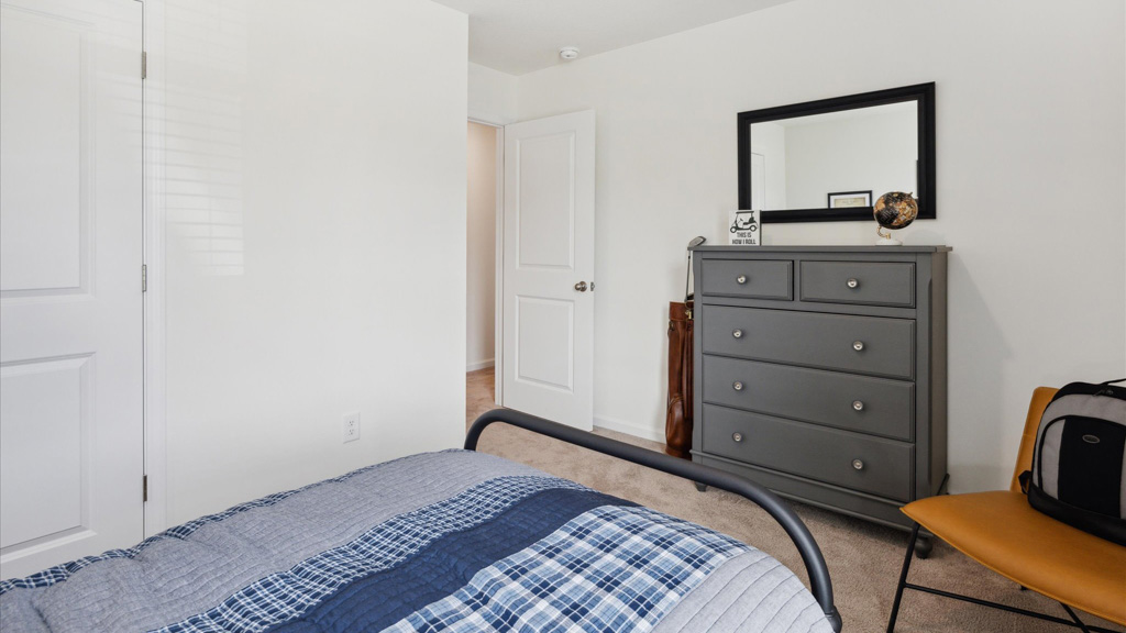 A view of a neatly organized bedroom featuring a gray dresser with a mirror above, a twin bed with blue plaid bedding in the foreground, and a white door leading to the hallway.