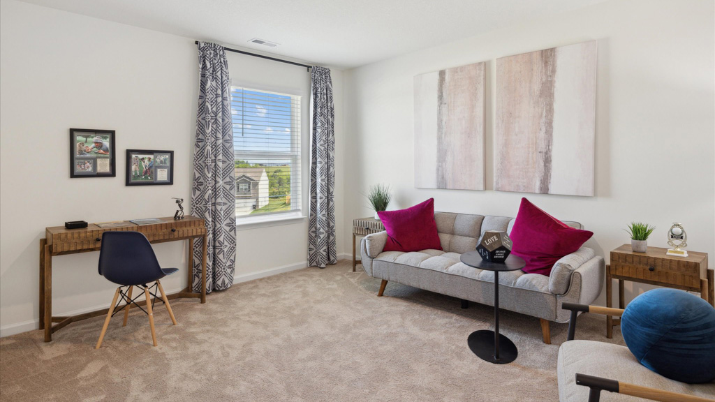 A bright and versatile room featuring a cozy loveseat with bold accent pillows, a small desk setup by the window, and neutral carpeting—ideal as a bedroom or flex space.
