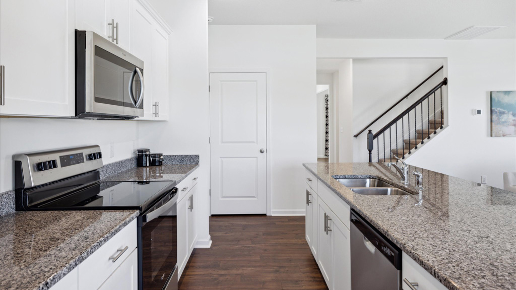 Contemporary kitchen with white cabinets, speckled countertops, stainless steel appliances, a double sink in the island, and a view of the adjacent staircase with dark railing and open layout beyond.