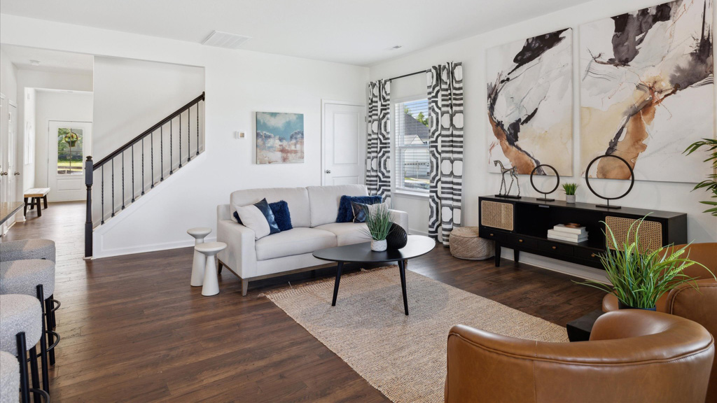 Contemporary living room with a light sofa, navy accent pillows, caramel leather chairs, abstract wall art, patterned curtains, and dark wood-look flooring, with a staircase and hallway visible in the background.