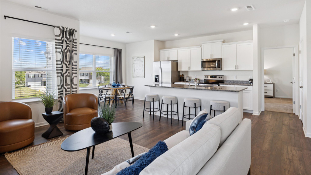 Open-concept living space featuring a modern kitchen with white cabinetry and stainless steel appliances, a large island with barstools, a cozy living area with a white sofa and caramel chairs, and a bright dining nook framed by large windows and patterned curtains.