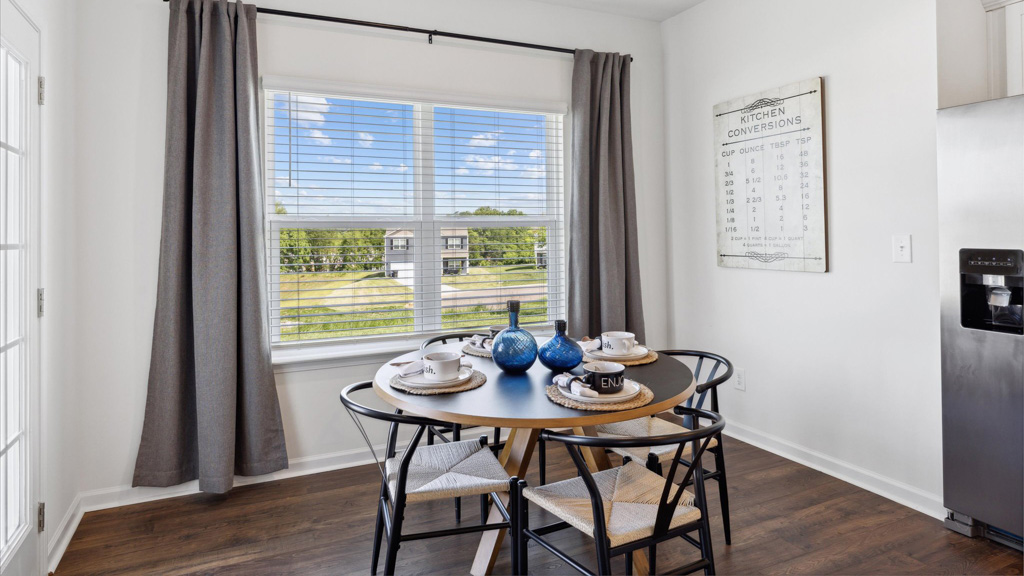 Bright breakfast nook with a round black dining table set for four, woven seat chairs, large window with gray curtains, and a wall-mounted kitchen conversions chart for added charm.