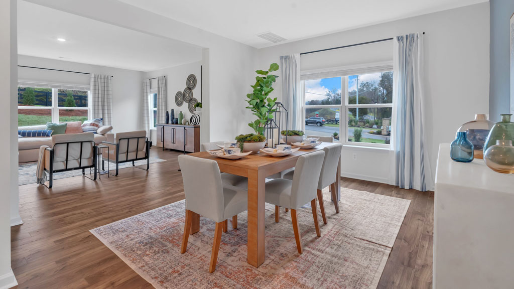 Open-concept layout featuring a dining area with a wooden table and six upholstered chairs, large windows with light curtains, and a view into a stylish living room with modern decor and natural light.