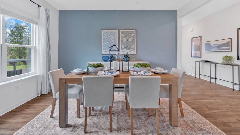 Modern dining room with a wooden table set for six, upholstered chairs, a blue accent wall with framed artwork, and large windows bringing in natural light.