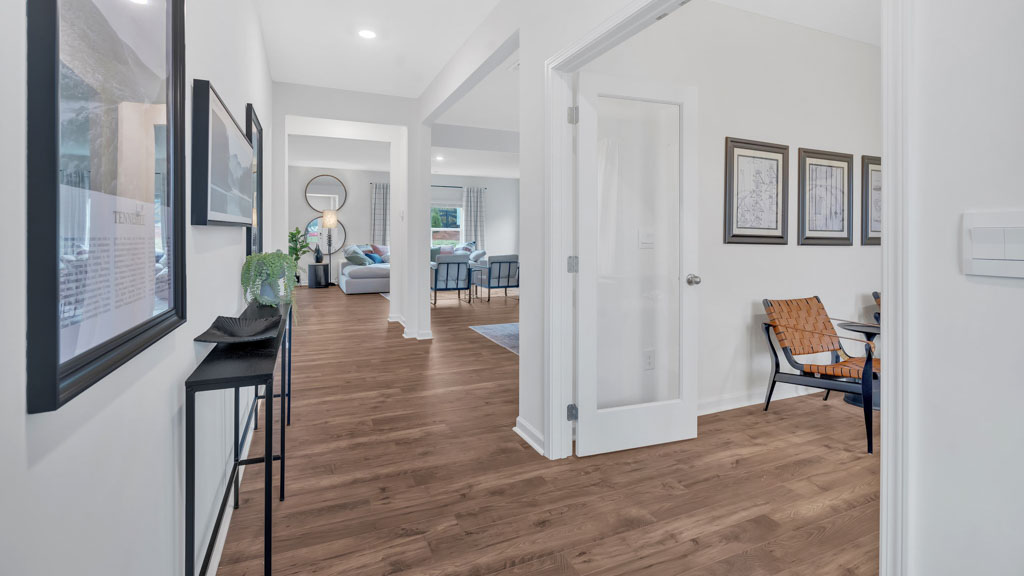 Front entrance hallway featuring warm wood flooring, wall art, a black console table with decor, and views into a bright, open-concept living space and adjacent flex room with glass door entry.