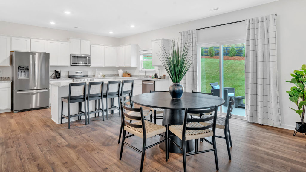 Modern open kitchen and dining area featuring white cabinetry, stainless steel appliances, a large island with bar seating, a round black dining table with woven chairs, and sliding glass doors leading to a patio and grassy backyard.