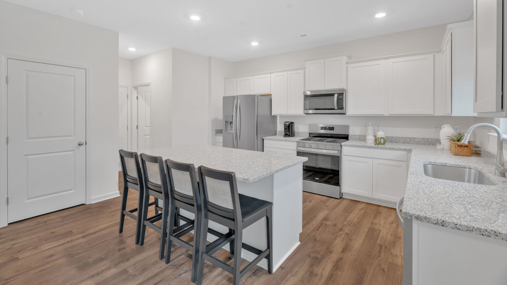 Modern kitchen with white cabinetry, stainless steel appliances, speckled countertops, a large center island with four barstools, and warm wood-style flooring throughout.