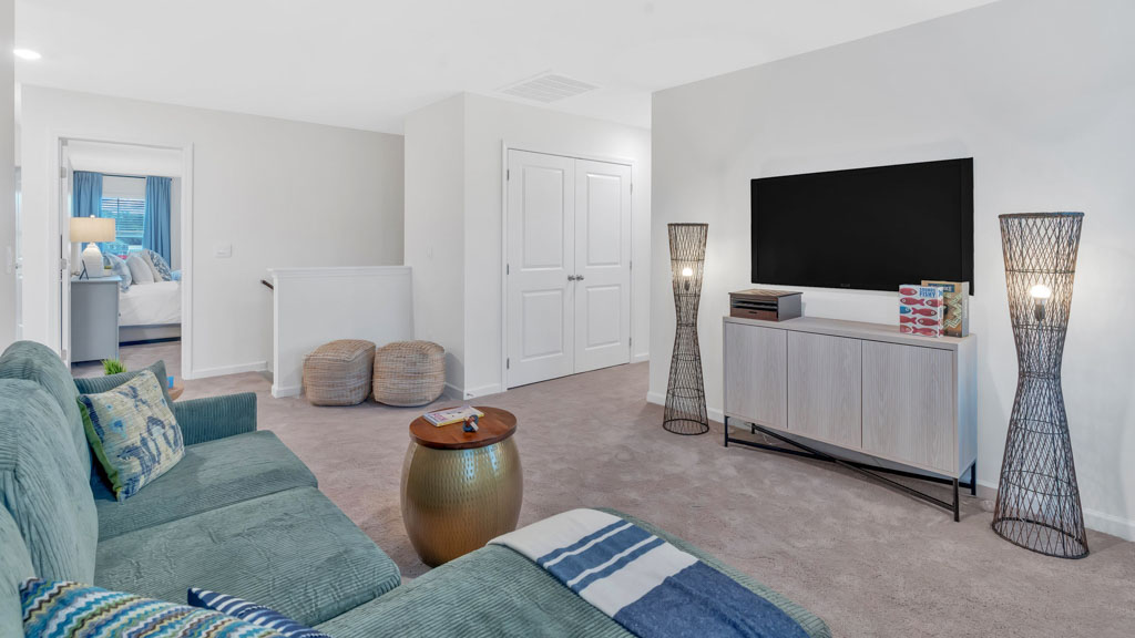 Upstairs loft area with a cozy green sectional, wall-mounted TV above a modern console, two woven floor lamps, soft carpet flooring, and a hallway view into a bedroom with a neatly made bed.