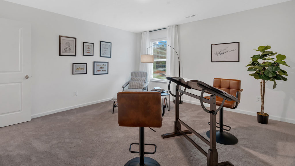 Stylish home office or study space featuring two industrial-style drafting desks with leather stools, a reading nook with a modern armchair and floor lamp, framed artwork on the walls, and a large window with white curtains.