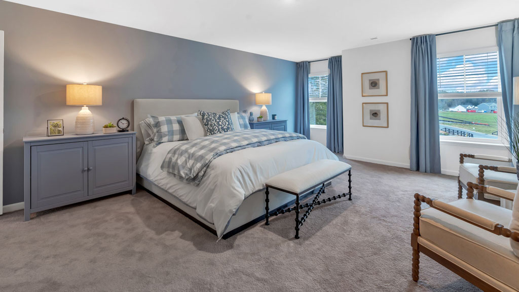 Elegant primary bedroom featuring a king-sized bed with blue plaid bedding, coordinating nightstands with lamps, a soft carpeted floor, two accent chairs, and large windows with blue drapes letting in natural light.