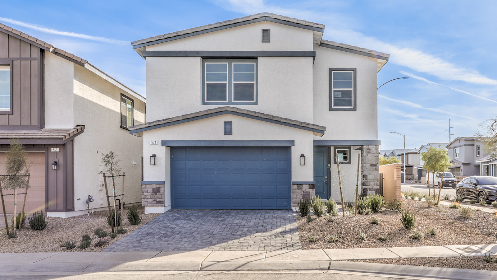 Our 2436 model home in the Symmetry Falls community with light grey and blue exterior paint, a flat tile roof, 2-car garage, front yard landscaping, and paver driveway and walkways.