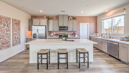 The Symmetry Falls 3000 Model Kitchen with Luxury Vinyl Plank flooring, featuring Whirlpool stainless steel appliances, a quartz countertop with a 4” backsplash, and a large center island with seating. The kitchen includes ample cabinetry, a walk-in pantry, and expansive windows that bring in natural light for a bright and functional cooking space.
