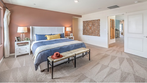 The Symmetry Falls 3000 Model Main Bedroom Suite with plush wall-to-wall carpet, large windows providing natural light, and an open layout designed for comfort and relaxation. The suite features an accent wall and direct access to a spacious ensuite bathroom and walk-in closet, creating a private retreat within the home.