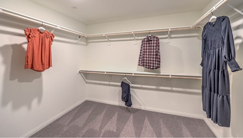 The Symmetry Falls 3000 Model Main Bedroom Walk-In Closet featuring wall-to-wall carpet, built-in dual-level hanging rods, and ample space for wardrobe storage and organization. The open layout offers easy access from the main bedroom suite bathroom, providing both convenience and functionality.