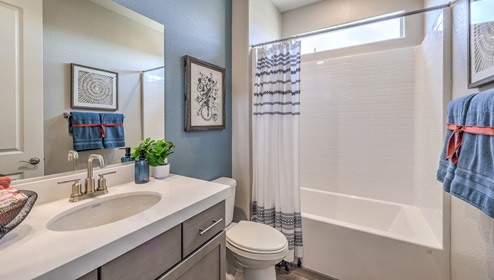 The second bathroom in our Heartland Manor 2754 Plan features luxury vinyl plank flooring, a vanity with quartz or granite countertops, and a combination shower and bathtub with a tiled surround. A large mirror enhances the space, while a high window allows natural light to brighten the room.