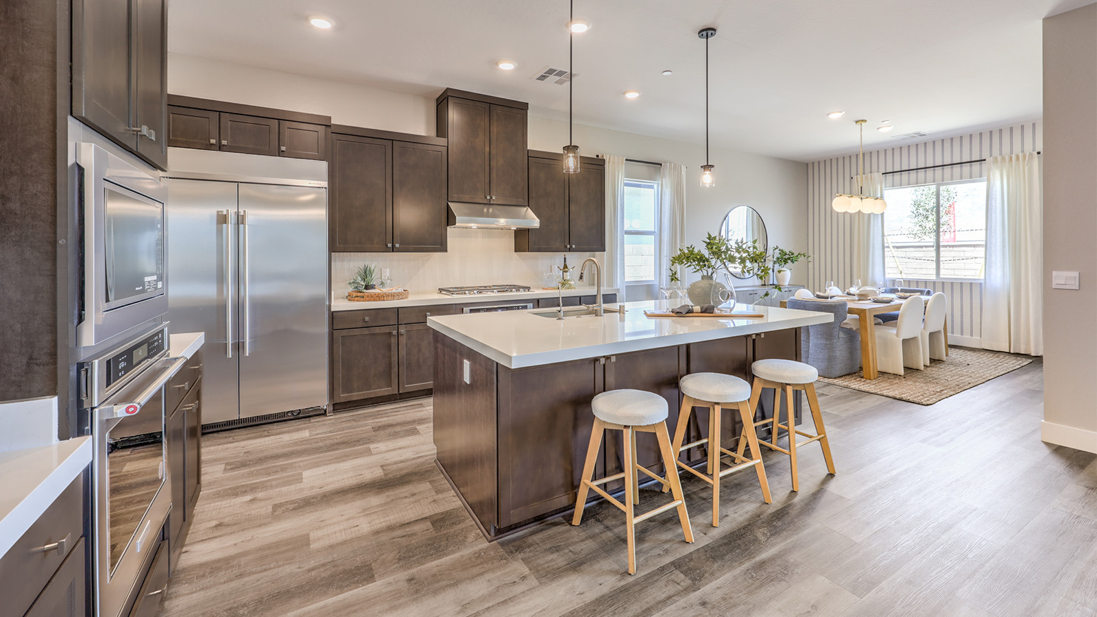 The kitchen in our Heartland Manor 2538 Plan features luxury vinyl plank flooring, an open-concept design, and a spacious layout. A large island with seating provides ample counter space, complemented by quartz or granite countertops with a 2¼-inch edge. Whirlpool stainless steel appliances include a five-burner cooktop, built-in ovens, a microwave, and a dishwasher. The space also features 42-inch upper cabinets for extra storage and a walk-in pantry for added convenience. Large windows bring in natural light, creating a bright and inviting atmosphere.