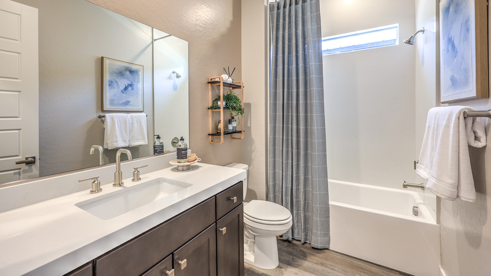 The second bathroom in our Heartland Manor 2538 Plan features luxury vinyl plank flooring, a vanity with quartz or granite countertops, and a combination shower and bathtub with a tiled surround. A large mirror enhances the space, while a high window allows natural light to brighten the room.