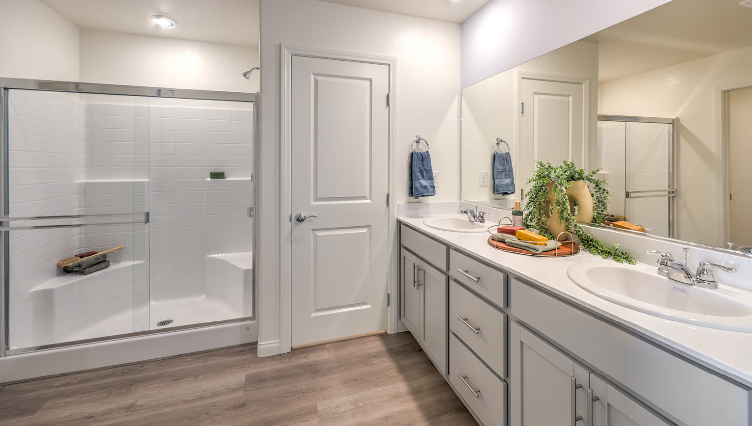 Attached to the Bedroom One Suite, our 1865 model bathroom with quartz/marble fusion countertops at the double Bath Vanity, a walk-in shower, mirror, and grey cabinets.