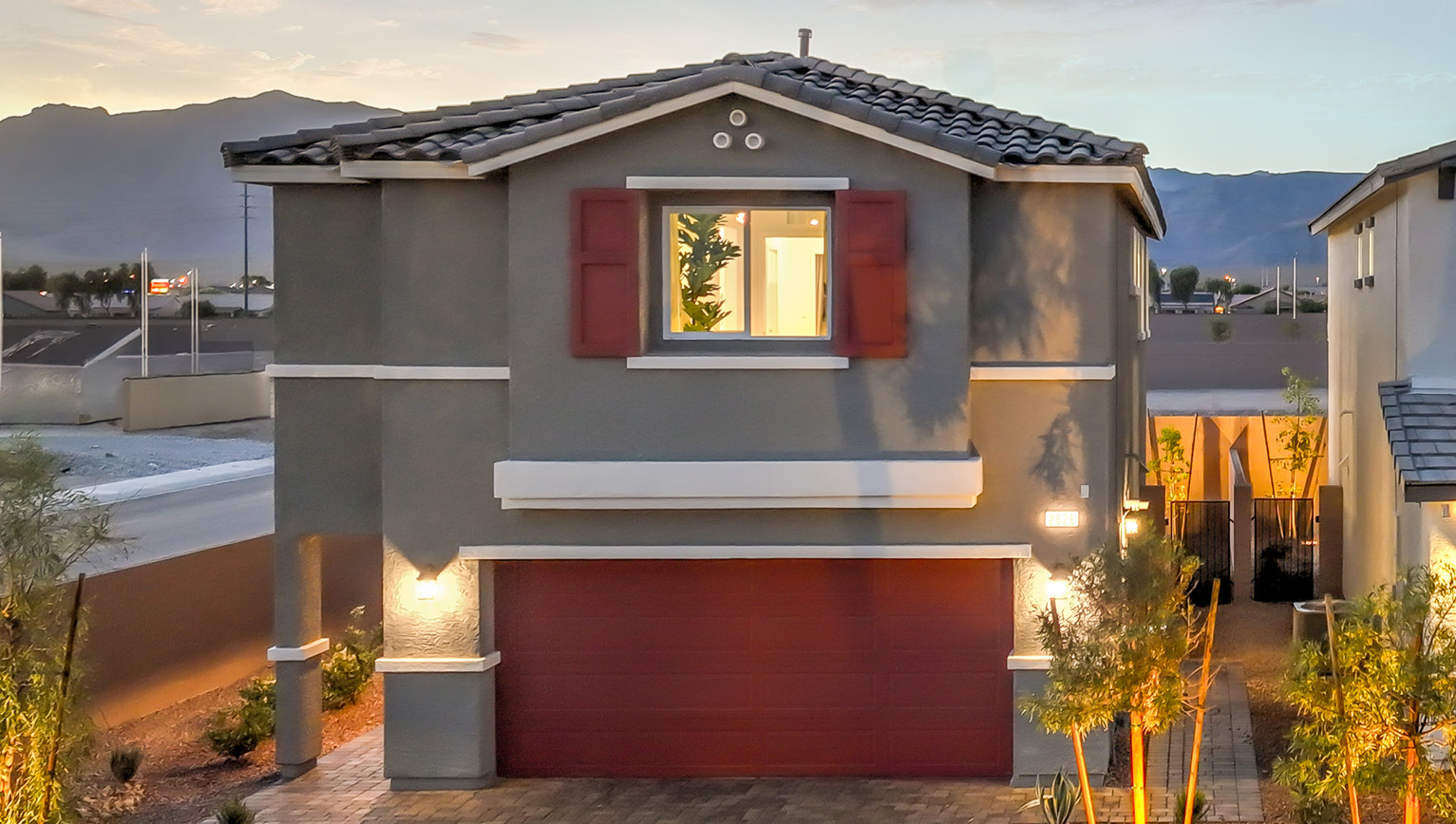 Our 1865 model home in the Kalea Bay community with red and grey exterior paint schemes, a flat tile roof, front yard landscaping, and paver walkways.