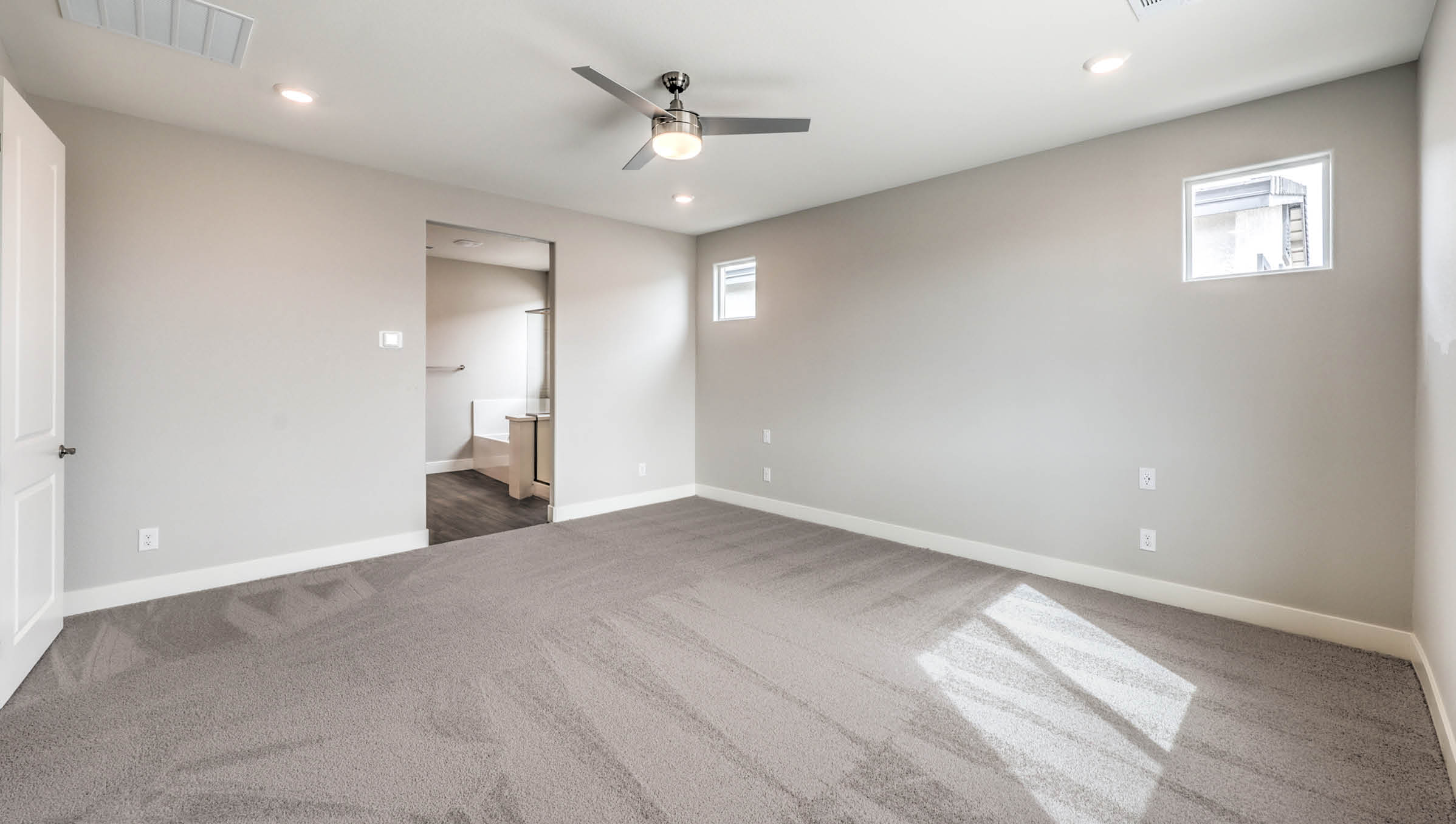 The Heartland Manor 2300 Plan Main Bedroom Suite featuring plush wall-to-wall carpet, multiple windows providing natural light, and a modern ceiling fan. This spacious layout offers a private retreat with direct access to the ensuite bathroom and walk-in closet, designed for comfort and relaxation.