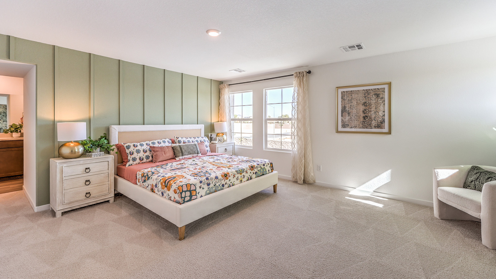 The Tropical Falls 3000 Model Main Bedroom Suite with plush wall-to-wall carpet, large windows providing natural light, and an open layout designed for comfort and relaxation. The suite features an accent wall and direct access to a spacious ensuite bathroom and walk-in closet, creating a private retreat within the home.