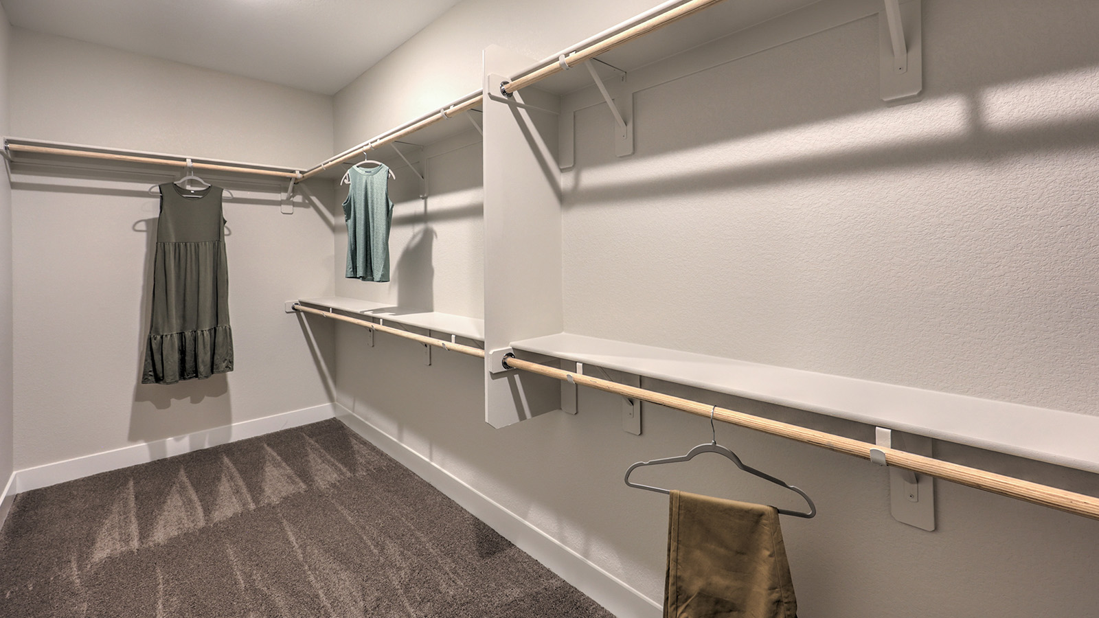 The Majestic Hills 2650 Plan Main Bedroom Suite Walk-In Closet featuring plush wall-to-wall carpet, built-in shelving, and double-level hanging rods. This spacious and well-lit closet offers ample storage and organization, conveniently accessible from the ensuite bathroom for everyday ease and comfort.