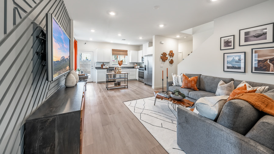 Open living space in Phoenix's model home, with an accent wall with carpentry details behind the wall-mounted TV and console table, a sectional sofa, live-edge coffee table, and a view into the adjoining kitchen and dining area.